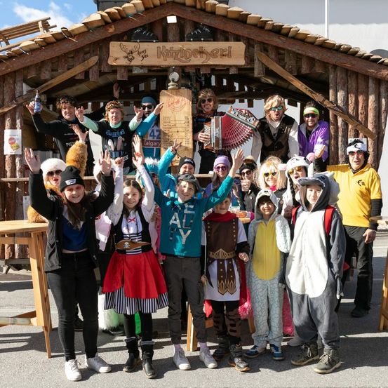 Eine Gruppe von Menschen in verschiedenen Kostümen posiert für ein Foto vor einem hölzernen Stand mit einem Schild, auf dem 'Kitzlocherl' steht. Einige von ihnen tragen Sonnenbrillen und Hüte. Hinter ihnen steht eine hölzerne Struktur mit einem Schild oben. In der Ferne gibt es Bäume und ein Gebäude.