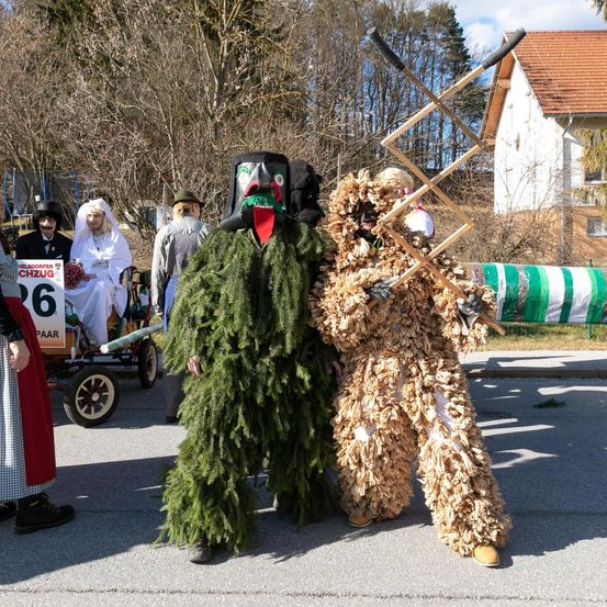 Zwei Personen in aufwendigen Kostümen stehen auf einer Straße, möglicherweise bei einer Parade. Einer trägt ein grünes Tannenkostüm, der andere ein braunes Laubkostüm. Hinter ihnen ist ein Radfahrzeug zu sehen, und ein Haus mit rotem Dach ist im Hintergrund.