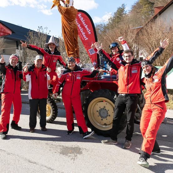 Eine Gruppe von Menschen in roten Overalls posiert für ein Foto vor einem Traktor vor einem Gebäude. Dahinter steht eine große Figur auf dem Traktor.