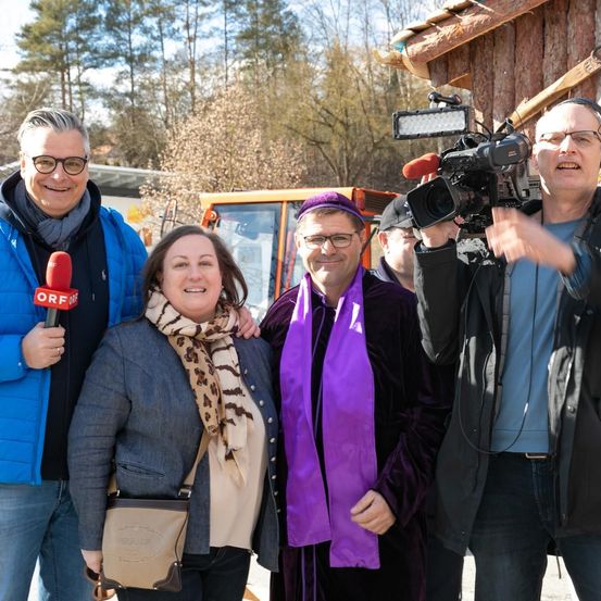 Drei Männer und eine Frau stehen zusammen draußen. Der Mann auf der linken Seite hält ein Mikrofon und der Mann auf der rechten Seite eine Kamera. Die Frau trägt einen Schal.