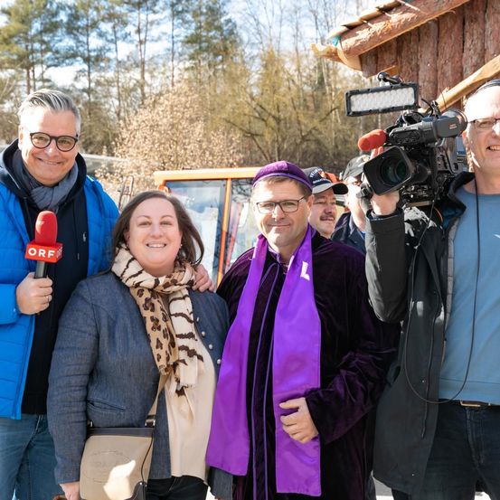 Eine Gruppe von Menschen steht draußen, einer mit einem Mikrofon, ein anderer mit einer Kamera. Hinter ihnen ein Fahrzeug und ein Holzgebäude. Bäume im Hintergrund.