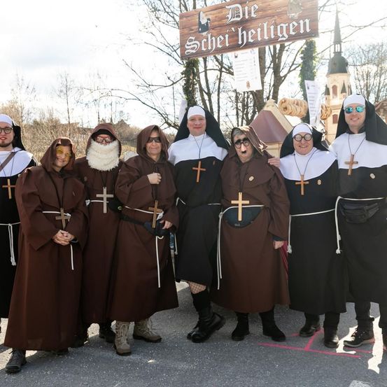 Eine Gruppe von Menschen, die als Nonnen und Mönche gekleidet sind, posiert für ein Foto vor einem Holzschild. Der Hintergrund besteht aus Bäumen und einem Gebäude.