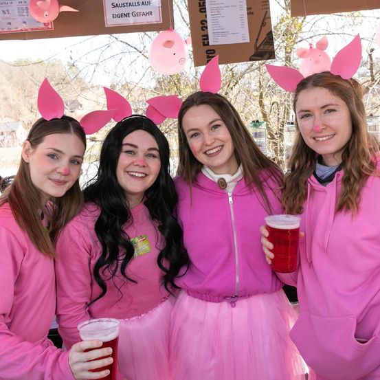 Vier Frauen mit rosa Schweineohren und rosa Outfits lächeln für die Kamera. Zwei von ihnen halten Gläser mit roten Getränken. Ein rosa Schweineballon und ein Schild hängen im Hintergrund.