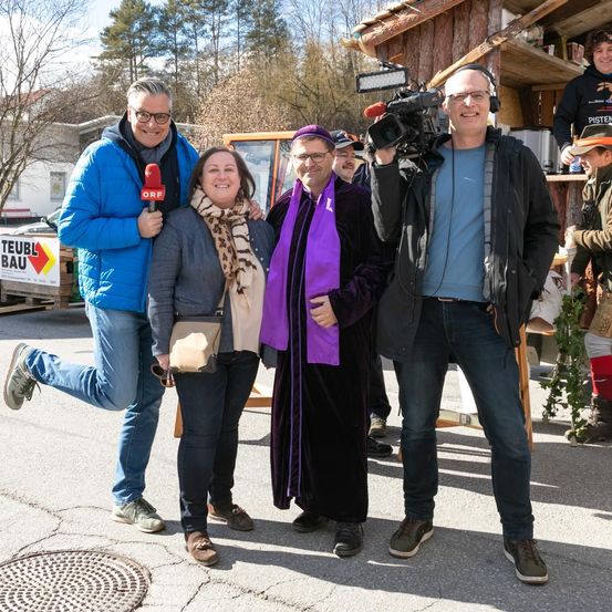 Eine Gruppe von Menschen steht im Freien, lächelt und posiert für ein Foto. Ein Mann hält ein Mikrofon und eine Kamera. Hinter ihnen steht ein Schild mit der Aufschrift Teubl Bau. Bäume und ein Haus sind im Hintergrund zu sehen.