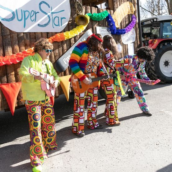 Eine Gruppe von Musikern in bunten Kostümen steht vor einem dekorierten Bus mit einem Traktor dahinter. Der Bus hat ein Schild, das Super Star steht.