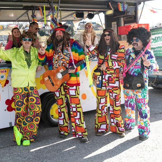Eine Gruppe von Menschen in farbenfrohen Outfits im Stil der 70er steht vor einem Food Truck und posiert für ein Foto. Sie haben Gitarren und andere Musikinstrumente.