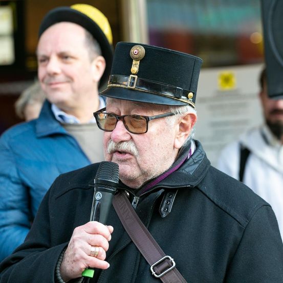 Ein älterer Mann mit einem Schnurrbart hält ein Mikrofon und spricht, trägt einen Hut mit einem dekorativen Abzeichen. Dahinter beobachten ein Mann in einem blauen Mantel und eine Frau mit einer Mütze.