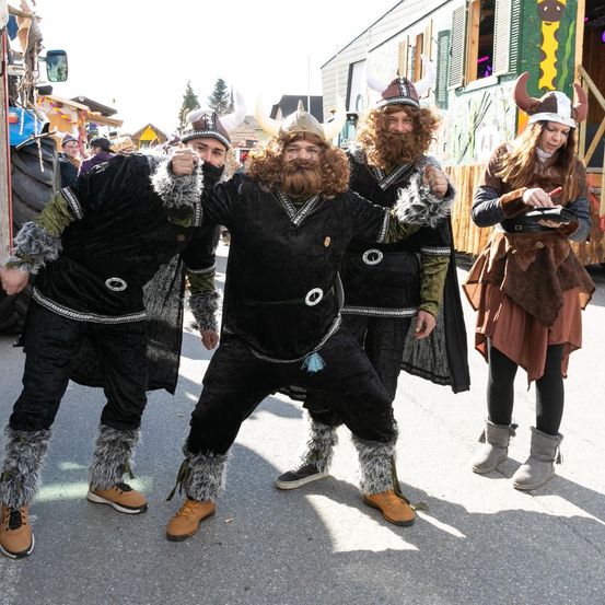 Vier Personen in Wikingerkostümen stehen auf der Straße, einer von ihnen gestikuliert. Hinter ihnen steht ein Traktor und ein geschmücktes Gebäude.