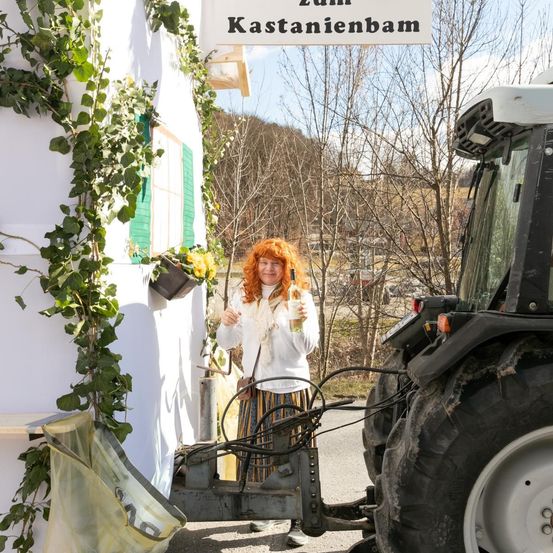 Eine Frau mit roten Haaren posiert neben einem Traktor und einem Gebäude mit dem Schild 'Zum Kastanienbaum'. Das Gebäude hat grüne Fensterläden und ist von Pflanzen umgeben.