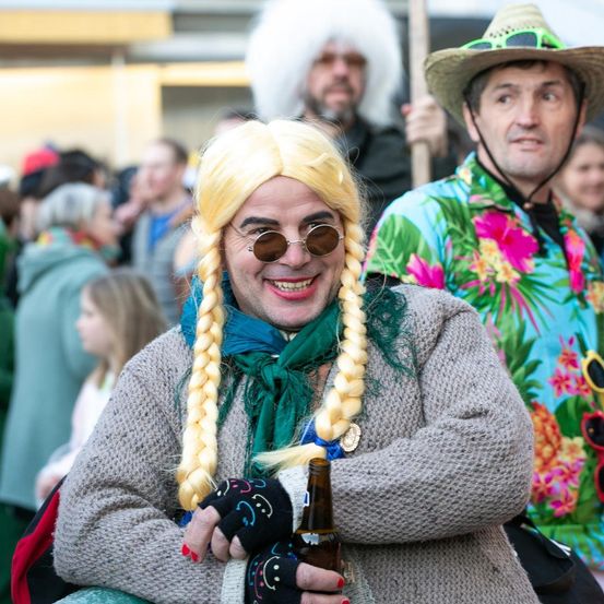 Ein Mann mit blonder Perücke und Sonnenbrille lächelt in die Kamera und hält ein Bier in der Hand. Hinter ihm beobachtet ein Mann mit Strohhut und Blumenhemd. Im Hintergrund sind Menschen in verschiedenen Kostümen gekleidet.