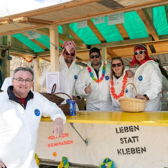 Fünf Personen in weißen Overalls posieren an einem Stand mit grünem Baldachin. Einer hat einen Schweinekopf auf. Ein Korb und Dosen stehen auf dem Tresen, der 'Leben statt Klebe' sagt.