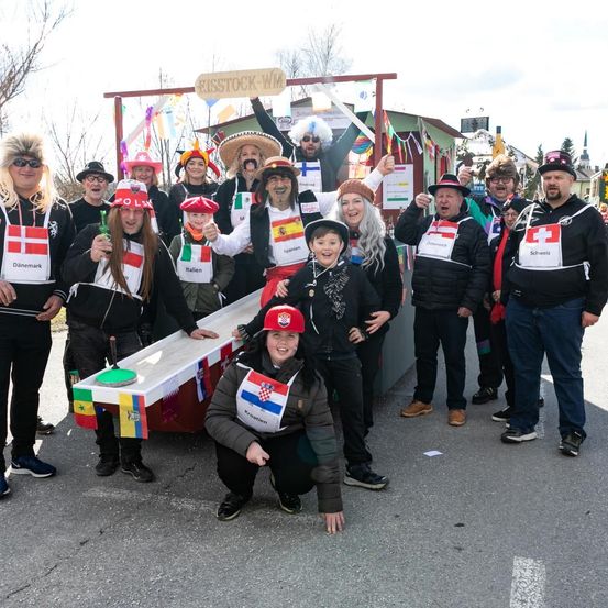 Eine Gruppe von Menschen in Kostümen, einige mit Flaggen, posiert für ein Foto auf einer Straße. Ein Schild im Hintergrund lautet 'MISSSTOCK-WIM'.