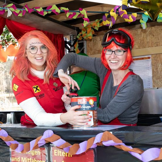 Zwei Frauen mit roten Haaren stehen hinter einem Tresen, eine trägt eine Schutzbrille und hält eine Dose Felix, die andere hat 'WAGEN' auf der Stirn geschrieben. Sie lächeln und scheinen sich auf einem Festival zu befinden.