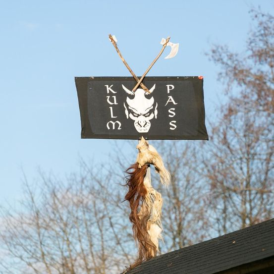 Ein schwarzes Banner mit weißen Buchstaben und einem weißen Schädel ist auf einem Dach montiert, mit zwei Äxten obenauf. Der Himmel ist klar und blau.