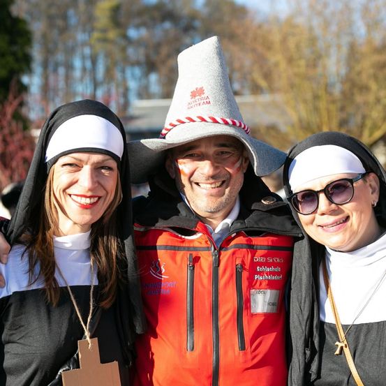 Drei Personen stehen zusammen in einer Außenumgebung. Zwei Frauen sind in Nonnenkostümen gekleidet, während ein Mann in einer roten Jacke mit Hut zwischen ihnen posiert.