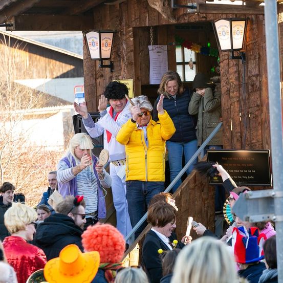 Eine Gruppe von Menschen in verschiedenen Kostümen steht vor einem Holzgebäude. Eine Person in einer gelben Jacke winkt der Menge zu. Eine Frau lächelt und macht ein Selfie. Ein Kind in einer grünen Jacke steht neben einer Person mit schwarzem Hut. Ein Schild ist an der Wand montiert.