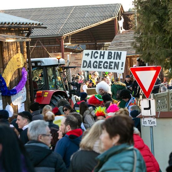 Eine Menschenmenge versammelt sich um einen Traktor. Einige halten Schilder hoch, eines davon steht 'ICH BIN DA GEGEN!' Ein rotes dreieckiges Schild befindet sich in der Nähe. Im Hintergrund sind Gebäude und Bäume zu sehen.