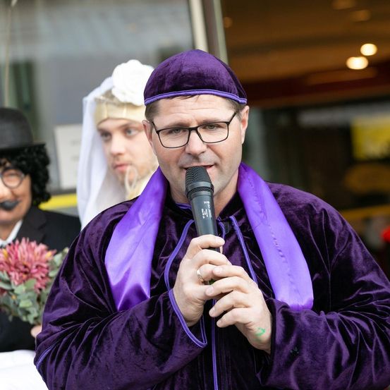 Ein Mann in einem lila Kostüm spricht in ein Mikrofon, während ein Mann in einem schwarzen Anzug und ein Mann in einem weißen Gewand hinter ihm stehen.