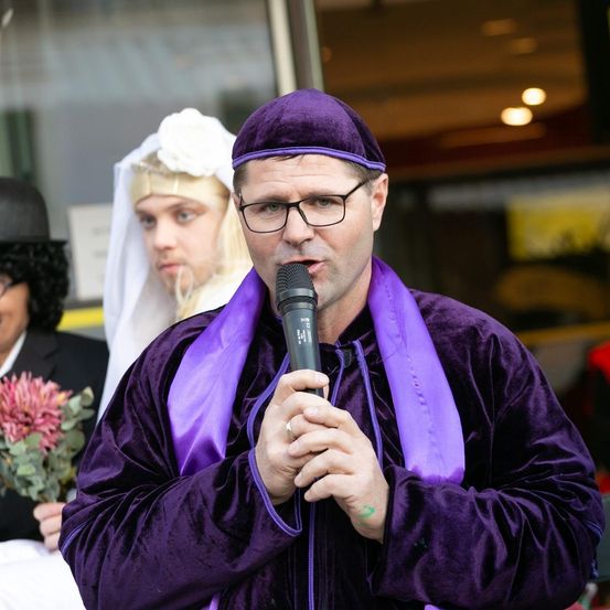 Ein Mann in einem lila Umhang spricht in ein Mikrofon, mit einer Frau in einem weißen Schleier und einem Mann in einem schwarzen Hut hinter ihm.