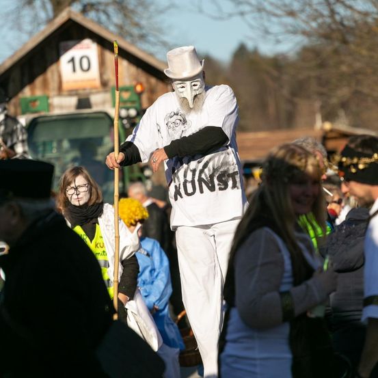 Ein Mann in einem weißen Kostüm mit Hut und Maske steht in einer Menschenmenge. Er hält einen Bambusstock. Die Menschen um ihn herum tragen verschiedene Kostüme. Ein Haus mit der Nummer 10 ist im Hintergrund zu sehen.