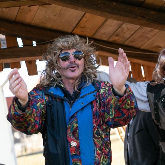 Ein Mann mit einer Perücke und Sonnenbrille, der eine bunte Jacke trägt, hebt seine Hände unter einer Holzkonstruktion. Eine weitere Person steht hinter ihm.