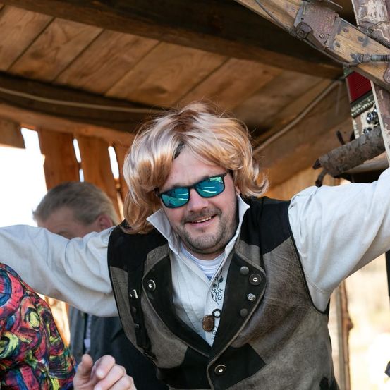 Ein Mann mit blondem Haar und Sonnenbrille lächelt in einer Holzstruktur. Ein weiterer Mann ist teilweise hinter ihm zu sehen.