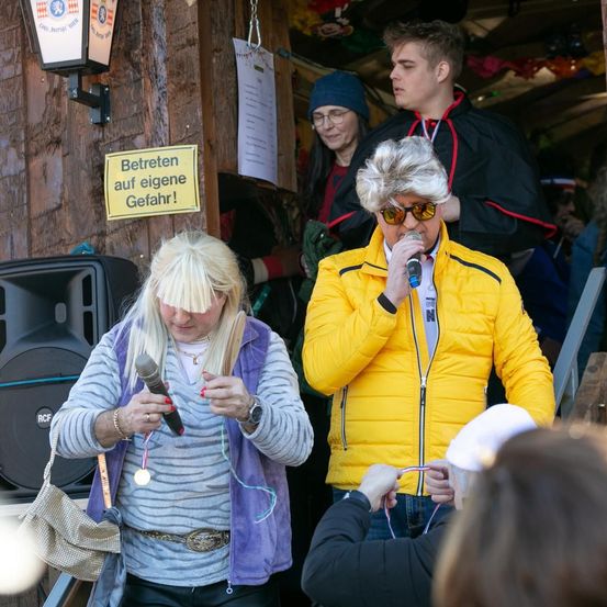 Zwei Frauen singen Karaoke und halten Mikrofone. Ein Schild an der Wand steht 'Betreten auf eigene Gefahr!' Ein Lautsprecher ist links, und Leute stehen hinter den Sängerinnen.