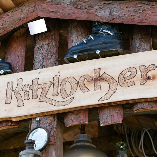 Ein Holzschild mit dem Wort Kitzlocher ist an einer Holzkonstruktion montiert. Der Schild ist in kursiver Schrift geschnitzt. Zwei schwarze Stiefel sind am Schild montiert. Die Konstruktion hat ein Holzdach mit einer kleinen Lampe.