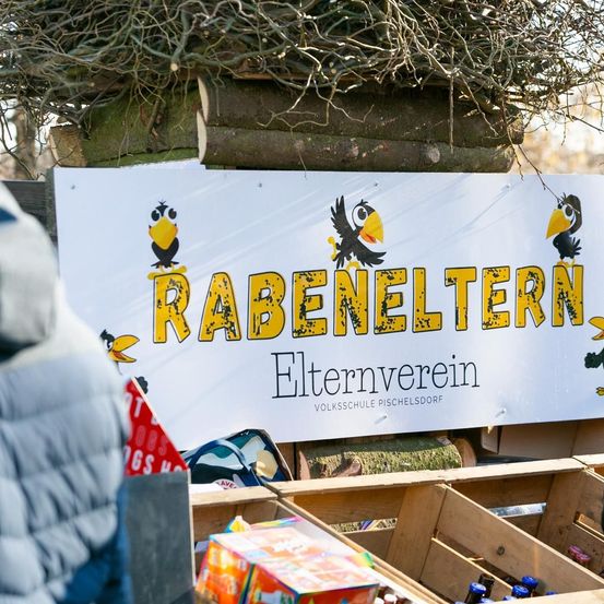 Ein weißes Schild mit dem Text 'RABENELTERN' und einer Cartoon-Bild von Vögeln ist auf einer Holzstruktur platziert. Unter dem Schild befinden sich einige Lagerboxen mit Gegenständen darin.