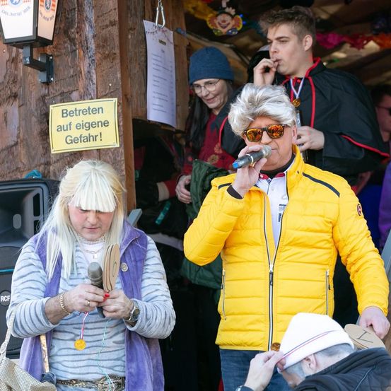 Ein Mann und eine Frau in einer Karnevalsumgebung, die Mikrofone halten. Ein gelbes Schild an der Wand steht 'Betreten auf eigene Gefahr!' oder 'Gefahren Sie auf eigene Gefahr!'.
