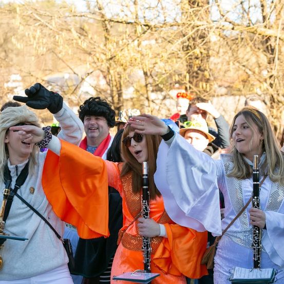 Eine Gruppe von Menschen in Kostümen führt eine musikalische Veranstaltung im Freien durch. Zwei Frauen halten Klarinetten und singen, während andere zusehen.