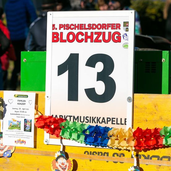 Ein weißes Schild mit rotem Text '5. PISCHELSDORFER BLOCHZUG' und der Nummer '13' ist zu sehen. Bunte Papierblumen hängen unten. Datum und Uhrzeit sind auf einem kleinen Flyer links angegeben.