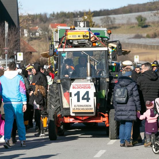 Ein geschmückter Traktor mit der Nummer 14 und den Worten 'BLOOCHUG' und 'ELTERNVEREIN VS PISCHELSDORF' fährt eine Straße entlang. Menschen in Kostümen beobachten.