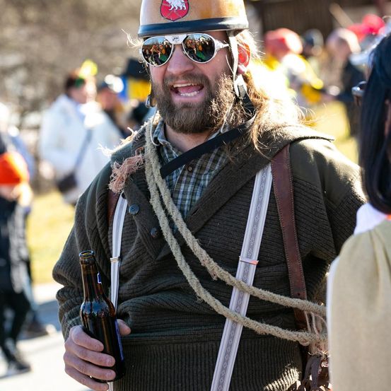 Ein Mann in einem gelben Helm und Sonnenbrille hält eine Flasche bei einer Outdoor-Veranstaltung, umgeben von Menschen in verschiedenen Kostümen.