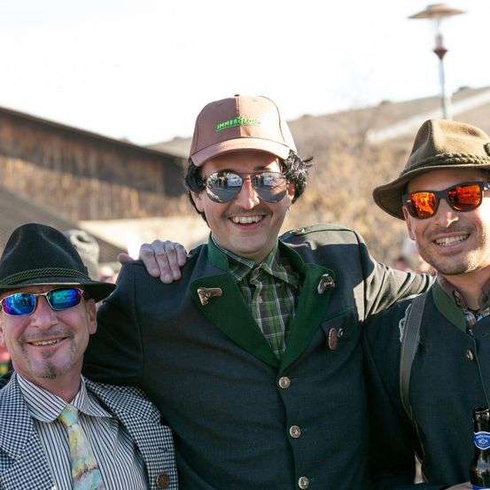 Drei Männer lächeln in traditioneller Kleidung bei einer Veranstaltung im Freien. Einer hält eine Bierflasche.