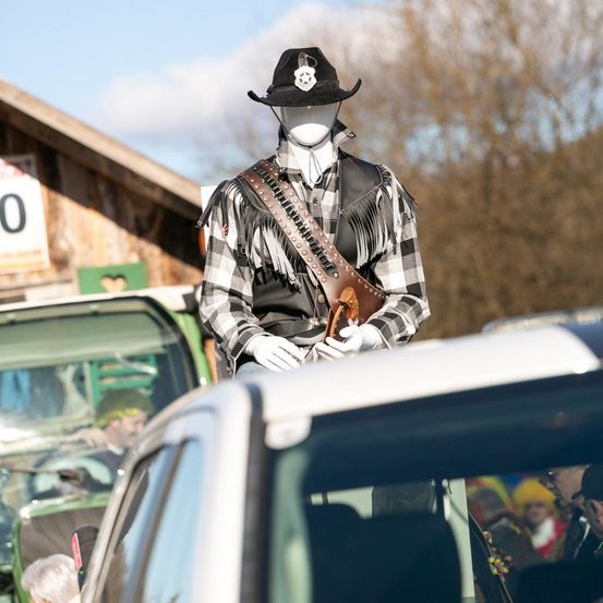 Eine Puppe in Cowboy-Kleidung steht auf einem Fahrzeug. Sie trägt einen schwarzen Hut und ein schwarz-weiß kariertes Langarmhemd. Im Hintergrund ist ein Gebäude mit einem Schild und Bäumen zu sehen.