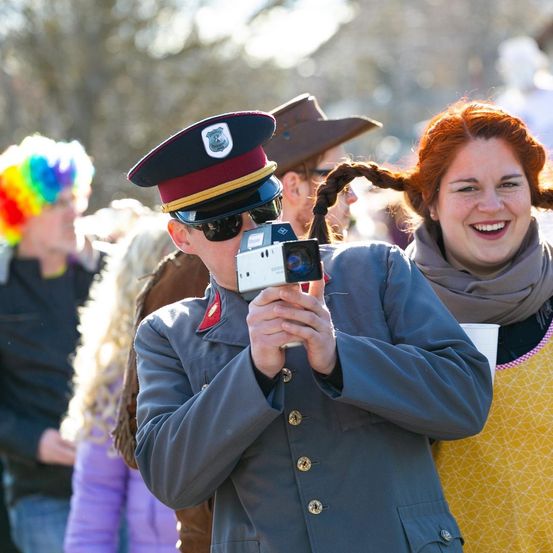 Ein Mann in Uniform hält eine Kamera und macht ein Foto einer lächelnden Frau mit langen Haaren. Sie sind von Menschen umgeben, einer trägt eine bunte Perücke.