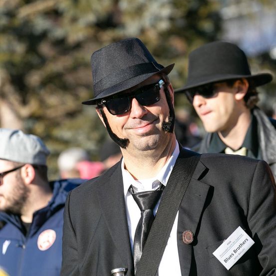 Ein Mann in einem schwarzen Anzug, Hut und Sonnenbrille lächelt, mit einem Namensschild Blues Brothers auf seiner Jacke. Hinter ihm blickt ein anderer Mann in einem schwarzen Hut und Sonnenbrille in die Kamera.
