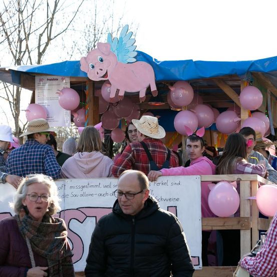 Eine Gruppe von Menschen versammelt sich vor einem Karnevalsstand. Der Stand zeigt ein rosa Schwein mit Flügeln oben, umgeben von rosa Luftballons. Einige Leute tragen Hüte, andere Brillen. Ein Schild mit rosa Buchstaben ist vorne angebracht.