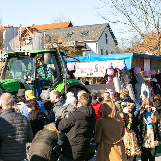 Eine Traktorparade mit Menschen in Kostümen und Luftballons. Einige tragen Blumenkränze. Ein Schild sagt 'Bauer sucht Sau'. Häuser und Bäume sind im Hintergrund.
