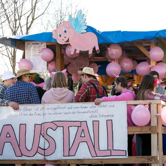 Eine Gruppe von Menschen steht um einen Karnevalsstand mit rosa Luftballons und einem Schwein als Maskottchen. Ein Schild steht 'AUSSTELL'. Die Leute tragen Hüte und Jacken.