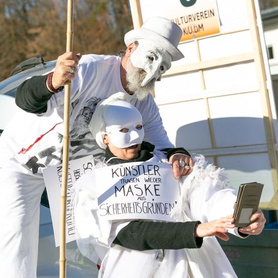 Zwei Personen in weißen Masken stehen draußen. Einer hält ein Schild mit der Aufschrift Künstler tragen Masken aus Sicherheitsgründen. Ein Banner im Hintergrund lautet 'Kulturverein KULM'.