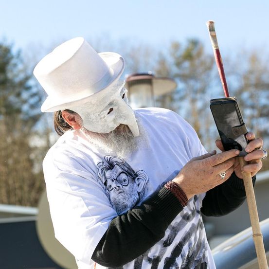 Eine Person mit weißer Maske und Hut, einem langen weißen Bart, hält ein Telefon und einen Pinsel in den Händen. Der Hintergrund zeigt Bäume und eine Straßenlampe.
