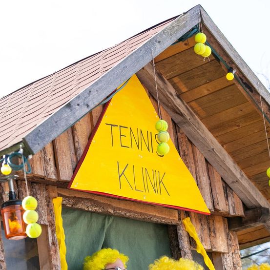 Ein hölzerne Struktur mit einem gelben dreieckigen Schild, das 'Tennis Klinik' besagt. Tennisbälle hängen vom Dach und eine gelbe gezackte Fahne ist an der Seite.
