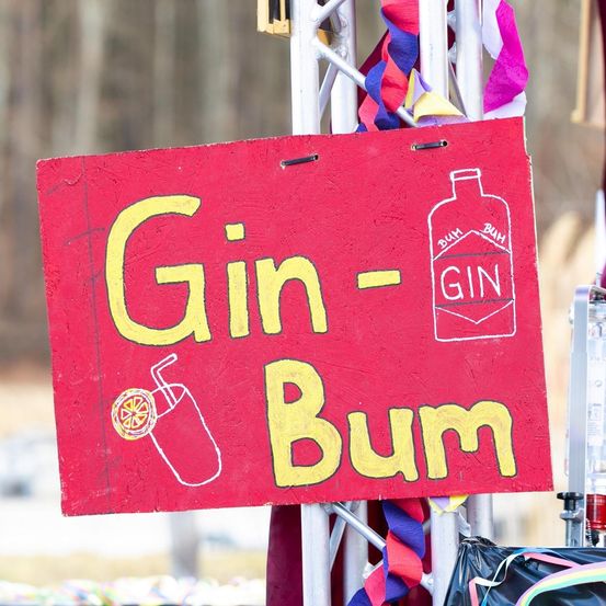Ein rotes Schild mit den Worten 'Gin-Bum' in Gelb geschrieben. Das Schild zeigt auch eine Zeichnung einer Ginflasche, auf der in Weiß 'GIN' steht. Das Schild ist an einem Metallpfahl befestigt.