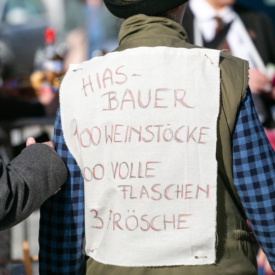 Eine Person trägt ein weißes Schild auf dem Rücken, auf dem 'HIAS BAUER 100 WEINSTOCKE 100 VOLLE FLACHEN 3 ROSCHE' steht. Im Hintergrund ist eine weitere Person teilweise zu sehen.