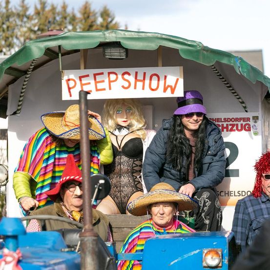Eine Gruppe von Menschen in Kostümen auf einem blauen Traktor. Eine Frau mit blonden Haaren ist in einem schwarzen Kleid. Ein Mann mit langen Haaren lächelt und sitzt an der Seite.
