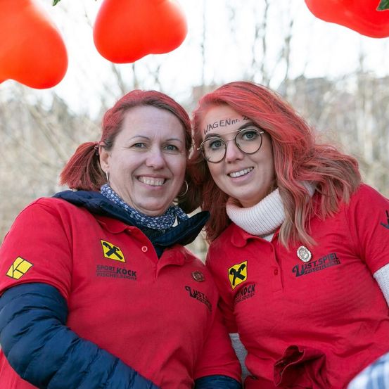 Zwei Frauen in roten T-Shirts mit gelben Logos posieren für ein Foto. Eine hat ein rotes Herz auf der Stirn. Sie lächeln vor einem unscharfen Hintergrund mit roten herzförmigen Ballons.
