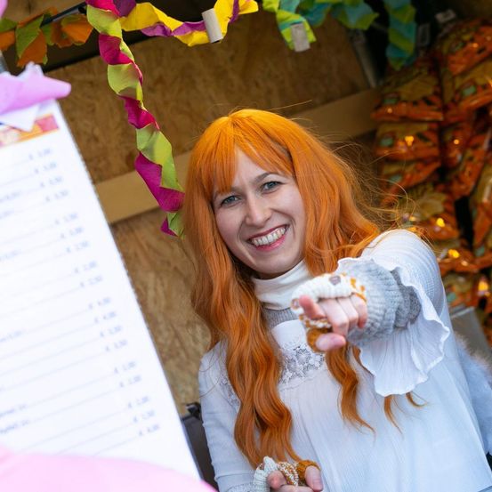 Eine Frau mit langen roten Haaren lächelt und zeigt auf ein Menü, während sie ein weißes Kleid und Handschuhe trägt. Sie steht neben einem Menü und Tüten mit Snacks.