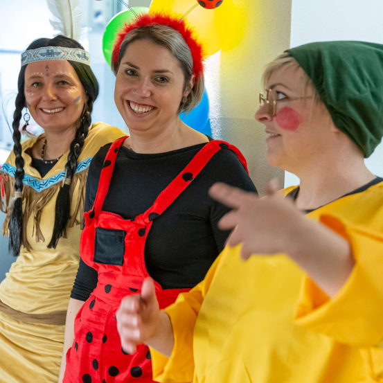 Drei Frauen in bunten Kostümen stehen eng beieinander und lächeln. Eine trägt eine rote Latzhose mit Punkten, eine andere einen grünen Hut und die dritte hat Gesichtsbemalung. Sie befinden sich in einem Raum mit Luftballons im Hintergrund.